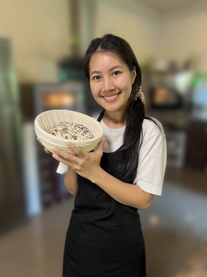 A smiling baker holding a cane proofing basket with a seeded sourdough