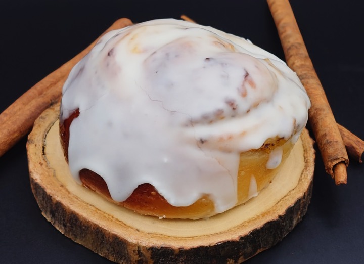 A large iced cinnamon roll on a wooden board with cinnamon sticks
